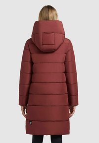 Rode gewatteerde winterjas met een grote kap, voorzien van horizontale stiksels en een klein logo-detail nabij de zoom. Gladde, matte textuur.