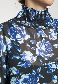 Black sheer blouse with ruffled smocked collar, featuring a floral pattern in shades of blue and white, with green accents on dark background.