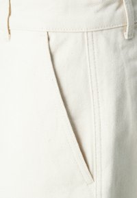 Pantaloni din bumbac bej cu o suprafață texturată. Prezintă buzunare laterale și cusături vizibile de-a lungul cusăturilor pentru un plus de detaliu.