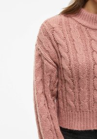 Roza pletena majica z visokim ovratnikom in rebrastim robom. Ima teksturirane vzorec in mehak, puhast videz. Posnetek rokava od blizu.