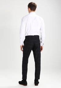Camicia bianca a maniche lunghe, vista posteriore, abbinata a pantaloni a scacchi neri. I pantaloni presentano una vestibilità slim e due tasche posteriori. Vengono indossate scarpe nere.