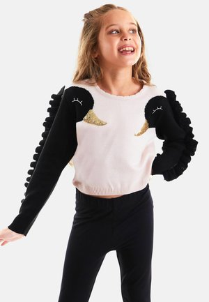 Weißer und schwarzer gestrickter Pullover mit Entenmuster, mit Paillettenakzenten und schwarzen, fransigen Ärmeln. Kombiniert mit schwarzen, figurbetonten Hosen.