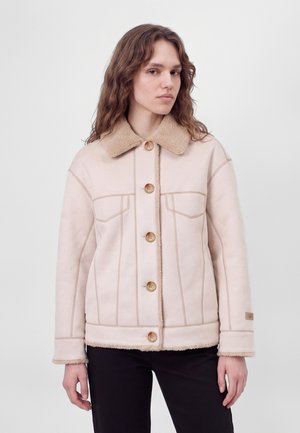 Una modelo de pelo largo y castaño lleva una chaqueta rosa pálido con cuello y puños de borreguito, cierre de botones delantero y paneles pespunteados.