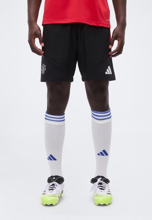 Athlète masculin portant un short noir de Manchester United avec logo Adidas, des chaussettes de football blanches avec des rayures bleues et des crampons avec des orteils vert néon.