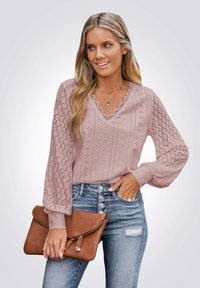 Femme blonde portant un blouse en dentelle lavande et un jean bleu, tenant une pochette marron, souriant et regardant sur le côté.