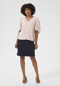 Blusa rosa chiaro a maniche corte con scollatura a V e chiusura con bottoni. Indossata con una gonna scura blu navy fino al ginocchio e sandali con tacco marroni.