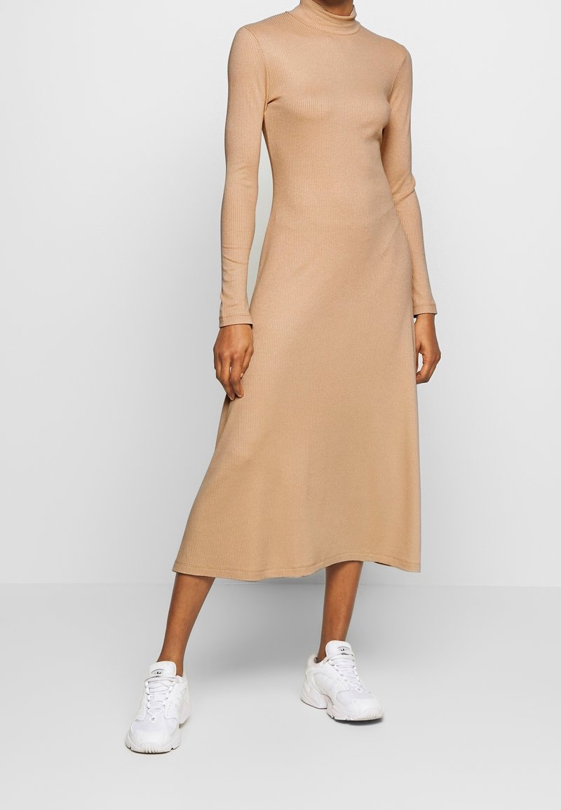 Beige ribbad långärmad klänning med hög halsringning, som faller till mitten av vaden. I kombination med vita sneakers. Mjuk textur och åtsittande design.