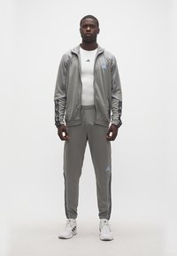 Άνδρας που στέκεται φορώντας γκρι φόρμα Adidas με λευκό μπλουζάκι Adidas και λευκά αθλητικά παπούτσια Adidas μπροστά σε απλό λευκό φόντο.