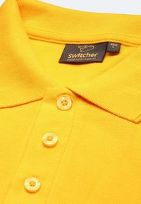 Gelbes Poloshirt aus strukturiertem Stoff, drei Knöpfe mit dem Markennamen und einem Etikett mit der Aufschrift "Switcher."