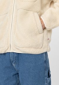 Kreemivärvi fleece-jakk, millel on pehme ja tekstureeritud pind. Omab tõmblukuga esiosa, küljetaskuid ja logo plaasterdusi allääres. Kanda siniste teksade peal.