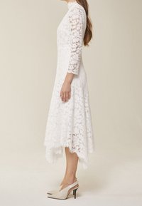 Abito di pizzo bianco con maniche a ¾ e scollatura alta, con orlo ondulato; abbinato a scarpe con tacco alto e cinturino beige.