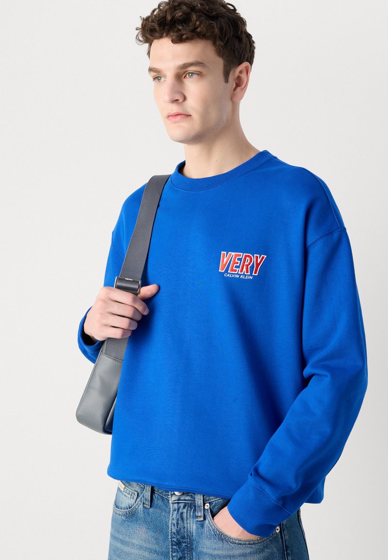 Sudadera de color azul real con mangas largas, cuello redondo y "VERY CALVIN KLEIN" bordado en rojo. Combinada con unos jeans de color azul claro y un bolso gris.