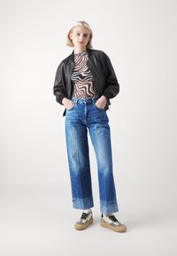 Chaqueta bomber negra, blusa de tela transparentes con estampado, vaqueros de tiro alto y piernas anchas en azul, y zapatillas con un diseño en blanco y negro.
