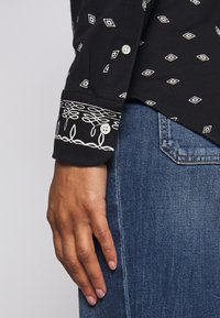 Svart långärmad skjorta med vita diamantmönster, med dekorerade manschetter. Bärs med blå jeans, som visar en avslappnad stil.