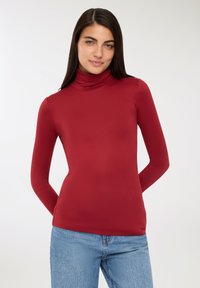 Czerwona bluzka z długim rękawem i golfem, wykonana z gładkiego materiału. Dopasowany krój z wysokim kołnierzem. W połączeniu z niebieskimi jeansami.