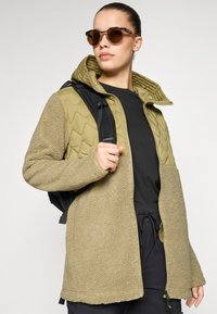 Olivenfarget jakke som kombinerer myk fleece og quiltet stoff, har hette, glidelås lukking og ekstra sidelommer. Svart ryggsekk inkludert.