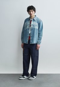 Ung mann står iført en denimjakke over en rød grafisk skjorte, mørke cargobukser og svart-hvite joggesko mot en ensfarget bakgrunn.