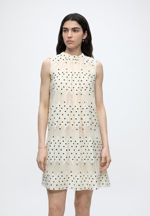 Femme aux cheveux foncés portant une robe sans manches crème à pois noirs, debout devant un fond blanc uni.
