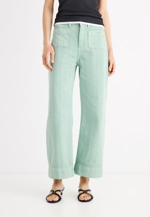 Osoba ubrana w jasnozielone szerokie spodnie z przednimi kieszeniami i czarne sandały na paski, stojąca na białym tle.