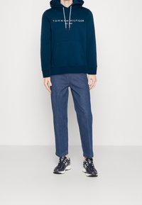 Felpa con cappuccio blu navy con tasca frontale, logo sul petto, abbinata a jeans azzurri e sneaker nere con dettagli bianchi.