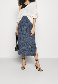 Übergroßer cremfarbener Strickpullover kombiniert mit einem blauen Midirock mit schwarz-abstractem Muster, seitlichem Schlitz und schwarzen Riemchen-Heels.