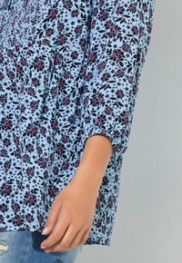 Niebieska kwiecista bluzka z czarnymi i czerwonymi wzorami, z marszczonym materiałem i długimi rękawami. Ręka spoczywa na jasnoniebieskich dżinsach.