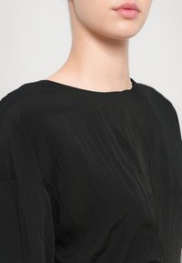 Jeune femme portant un haut noir texturé avec un col rond, vue du cou aux épaules sur un fond clair.