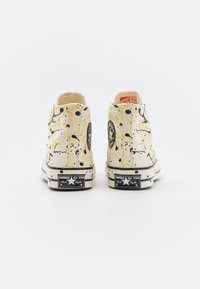 Tênis de cano alto em lona com base branca e design salpicado em amarelo e preto, detalhes em preto e um logo clássico da Converse na lateral.