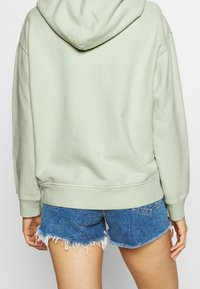 Svetle zelená mikina s kapucí, volného střihu, s žebrovaným lemem a manžetami, v kombinaci s modrými džínovými kraťasy. Viditelné detaily texturované tkaniny.