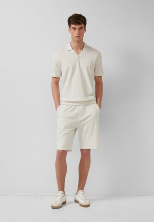 Młody mężczyzna stojący z rękami w kieszeniach, ubrany w beżową koszulkę polo z krótkim rękawem, pasujące spodenki i białe trampki na jednolitym tle.