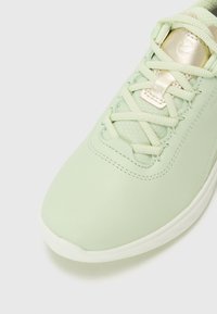 Sapatilha de couro verde claro com uma textura lisa, apresentando atacadores em creme e um detalhe metálico dourado na língua. Design de bico arredondado.