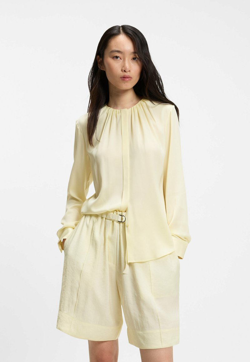 Femme aux longs cheveux foncés portant un chemisier ample jaune pâle et un short assorti avec ceinture, debout les mains dans les poches sur fond blanc.