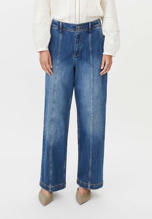 Person trägt hoch taillierte, weit geschnittene blaue Jeans mit vertikalen Nähten und beigen spitzen Schuhen, kombiniert mit einer weißen Spitzenbluse.