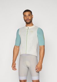 Maillot de cyclisme en tissu crème et canard, à manches courtes, avec une fermeture éclair à l'avant et une rayure multicolore, associé à un short de cyclisme gris.