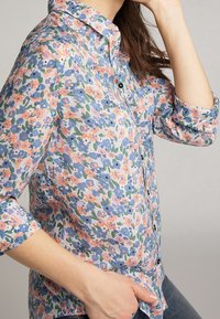 Camicia a maniche lunghe con bottoni, in tessuto leggero, con fiori blu e arancioni su uno sfondo chiaro, maniche arrotolate ai polsini.