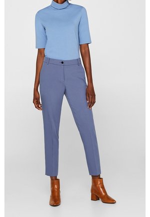 Kobieta w jasno niebieskim krótkim golfie z krótkim rękawem, granatowych dopasowanych spodniach i brązowych botkach za kostkę, stojąca na jednolitym tle.