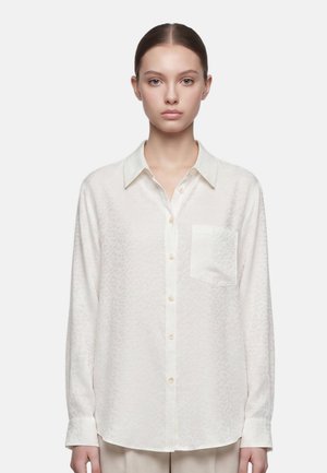Giovane donna con i capelli legati che indossa una camicia beige chiaro a maniche lunghe con tasca sul petto, in piedi davanti a uno sfondo semplice.