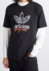 Černé bavlněné tričko s lakovaným stříbrným a oranžovým logem Adidas, krátkými rukávy a kulatým výstřihem, vrstvené přes bílé tričko s dlouhým rukávem.