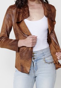 Hnědý kožený blazer s volným límcem, hladkým povrchem a dlouhými rukávy. V kombinaci se světle modrými džíny s vysokým pasem a ozdobnými knoflíky.