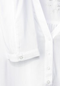 Camicia bianca con maniche corte, realizzata in tessuto leggero. Presenta una patta con bottoni simili a perle e un dettaglio plissettato sulla schiena.
