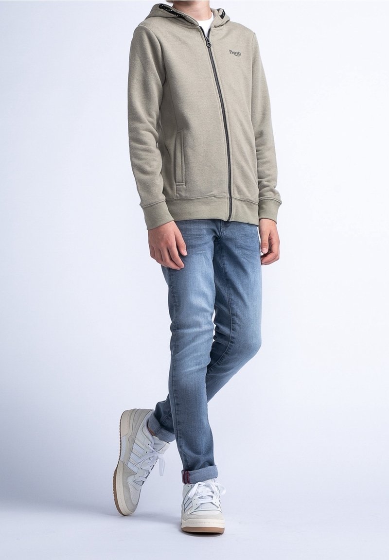 Haut de survêtement zippé gris clair avec capuche à cordon, associé à un jean bleu clair et des baskets montantes blanches avec des accents rouges.