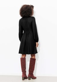 Robe noire à manches longues avec une taille froncée, associée à des bottes en daim bordeaux montantes jusqu'au genou avec un talon épais. Fond blanc uni.