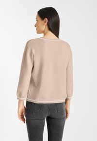 Vrouw met lang donker haar, gekleed in een beige trui met witte accenten en donkergrijze jeans, die met de rug naar de camera staat tegen een witte achtergrond.