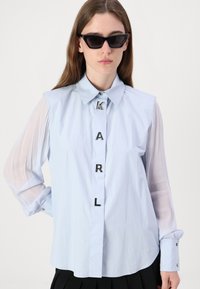 Camicia azzurro chiaro con maniche trasparenti, patta con bottoni caratterizzata da lettere argento K, A, R, L. Polsini con bottoni e dettaglio dell'orlo curvato.