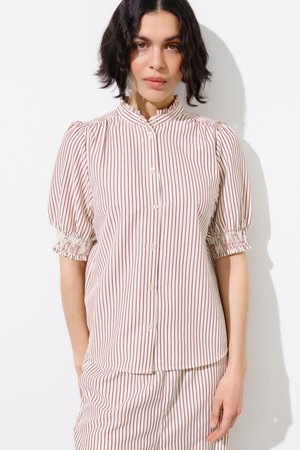 Lühikeste tumedate juustega naine kannab valget nööpidega pluusi ja sobivaid pükse, millel on õhukesed vertikaalsed punased triibud, seisab valgel taustal.