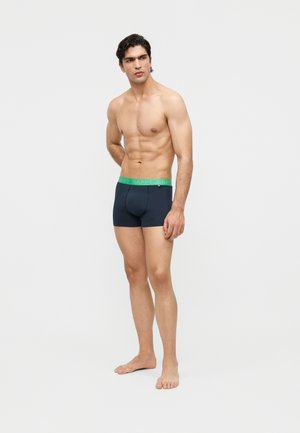 Mężczyzna bez koszulki stojący boso, ubrany w ciemne bokserki z zielonym pasem z napisem "HAPPY", patrzący na bok.