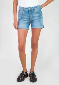 Shorts de mezclilla en azul claro con puños enrollados, que cuentan con bolsillos frontales y un cierre con cremallera, combinados con sandalias negras de tiras.