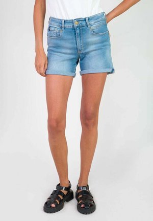 Shorts de mezclilla en azul claro con puños enrollados, que cuentan con bolsillos frontales y un cierre con cremallera, combinados con sandalias negras de tiras.