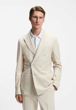 Homme portant un blazer crème côtelé croisé sur une chemise blanche à boutons et un pantalon clair, debout avec une main dans la poche.