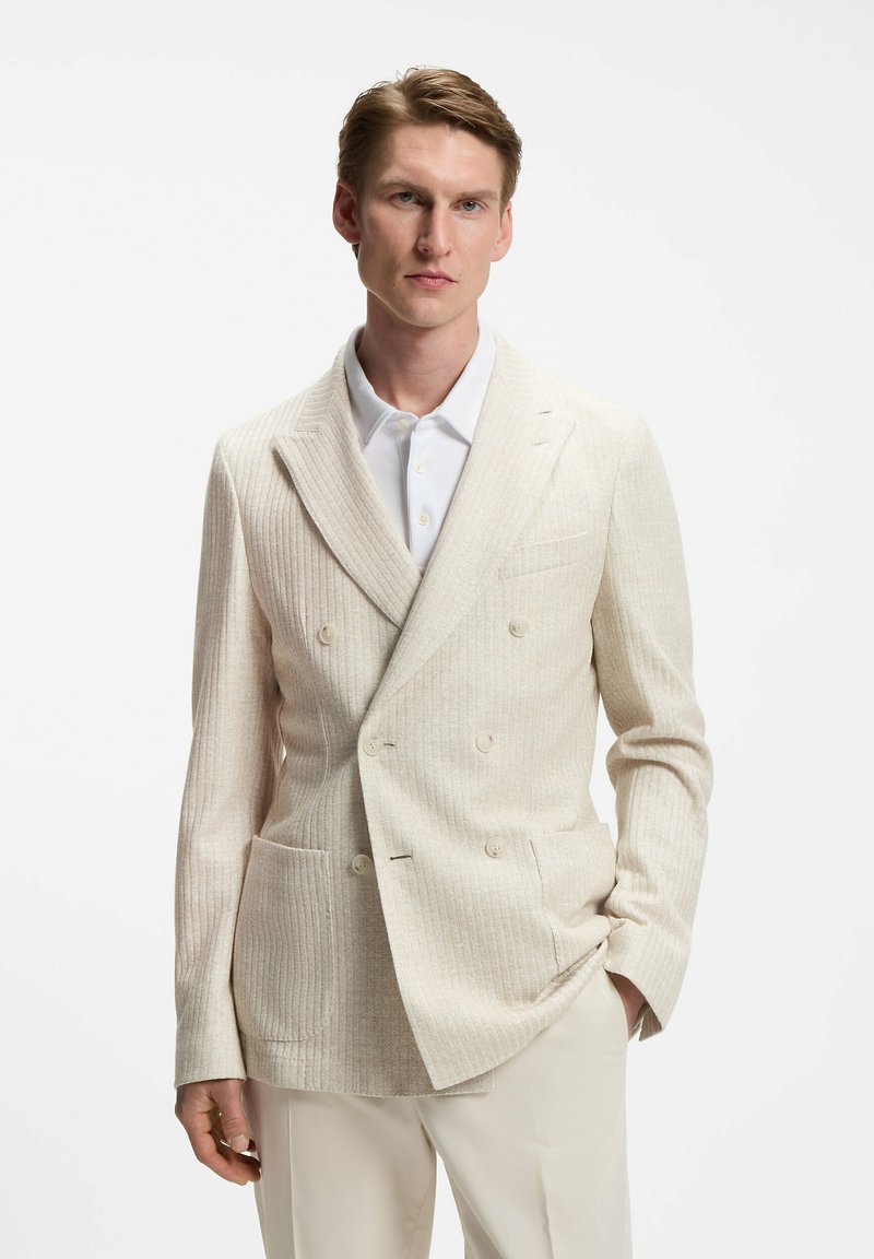 Homme portant un blazer crème côtelé croisé sur une chemise blanche à boutons et un pantalon clair, debout avec une main dans la poche.